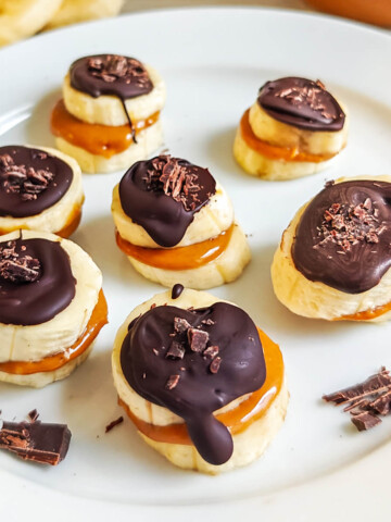 Bouchées de rondelles de bananes garnies de beurre de cacahuète crémeux et nappées de chocolat noir fondu, parsemées de copeaux de cacao sur une assiette blanche.