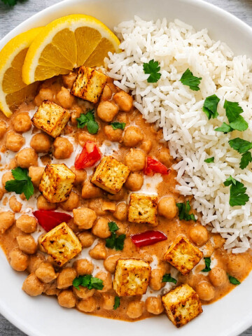 Un bol blanc contenant un curry de pois chiches onctueux et carrés de tofu au lait de coco et à la tomate, servi avec du riz complet, décoré de tranches de citron jaune et de coriandre fraîche.