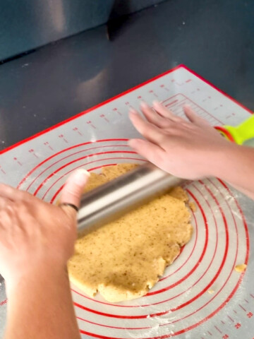 Vue de dessus montrant des mains étalant une pâte à tarte maison avec un rouleau en inox sur un tapis de cuisson gradué.