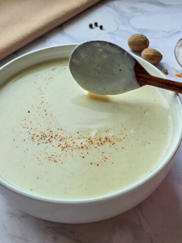 Un bol contenant de la sauce béchamel fait maison, sur fond marbré blanc, noix de muscade, avec une cuillère en bois.