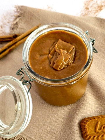 Pot de pâte à tartiner aux spéculoos maison onctueuse avec un biscuit Lotus et des bâtons de cannelle sur une nappe en lin.