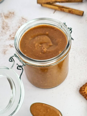 Bocal en verre ouvert de pâte à tartiner aux spéculoos vegan onctueuse avec une cuillère remplie au premier plan, biscuits Lotus et cannelle sur fond blanc.