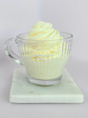 Une tasse en verre remplie de crème chantilly maison ferme et onctueuse, pochée en dôme avec des rainures bien nettes, posée sur un socle en marbre blanc.