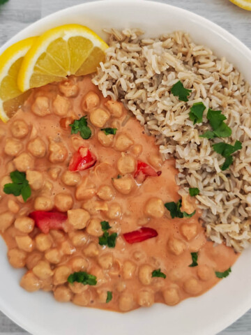 Un bol blanc contenant un curry de pois chiches onctueux au lait de coco et à la tomate, servi avec du riz complet, décoré de citron et de coriandre fraîche.