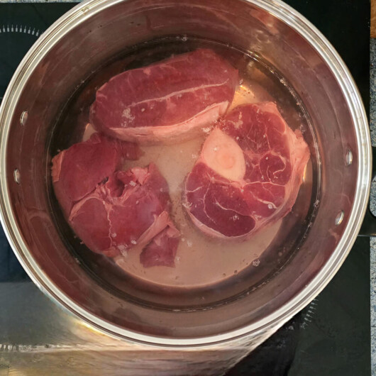 Trois gros morceaux de viande de bœuf, dont un avec os à moelle, immergés dans une casserole d'eau au début de la cuisson.