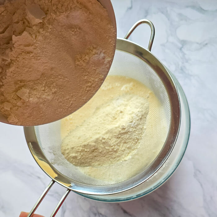 Ajouter-farine-tamisee-molly-cake Farine tamisée au-dessus d'un bol de préparation pour Molly Cake afin d'éviter les grumeaux et garantir une texture légère.