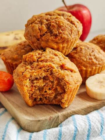 Muffins moelleux aux carottes, pommes et bananes servis sur une planche en bois. Un muffin est croqué pour montrer sa texture aérée à l'intérieur. En arrière-plan, les ingrédients frais sont visibles : une pomme rouge, une banane, des rondelles de banane et une carotte entière