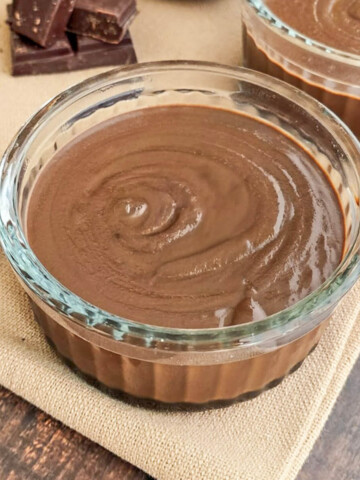 Deux ramequins en verre remplis de crème dessert onctueuse au chocolat et lait concentré, disposés sur une serviette en lin avec des carrés de chocolat noir et un pichet en céramique en arrière-plan.