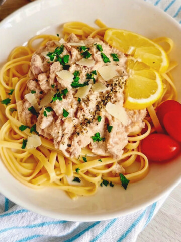Une vue de dessus d'une assiette de pâtes linguines en sauce crémeuse au thon, richement garnie de copeaux de parmesan, de persil frais haché et d'herbes séchées. Le plat est présenté avec deux tranches de citron et quelques tomates cerises sur le côté, sur un fond texturé.