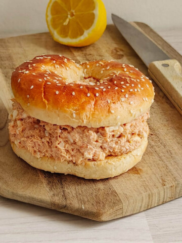Photo d'un bagel entier au thon crémeux, recouvert de graines de sésame dorées, présenté sur une planche à découper en bois. En arrière-plan, une assiette de salade verte (mâche), un demi-citron jaune et un couteau à manche en bois. La texture de la garniture au thon est bien visible.