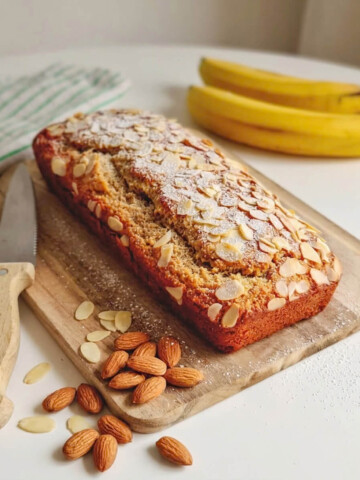 Un banana bread facon frangipane doré, parsemé d'amandes effilées sur le dessus, présenté sur une planche à découper en bois avec un couteau et deux bananes entières en arrière-plan.