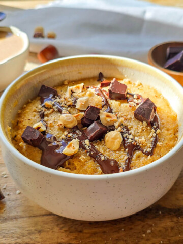 Un mug cake praliné chocolat noir dans un bol blanc, nappé de chocolat fondu, d'éclats de chocolat noir et de noisettes entières et concassées. Le bol repose sur une planche en bois à côté de carreaux de chocolat et de noisettes éparpillés.