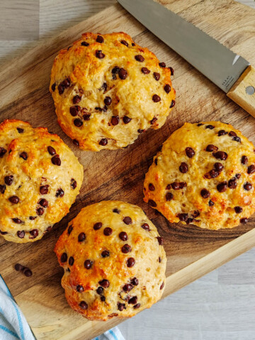 Photographie aérienne de quatre briochettes pépites de chocolat dorées et moelleuses, disposées en carré sur une planche à découper en bois rustique. Une lame de couteau est posée en diagonale sur la planche et un linge rayé bleu et blanc est visible sur le côté.