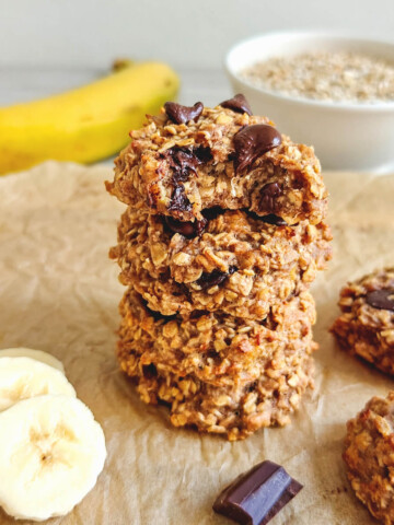 Une pile de trois cookies moelleux aux flocons d'avoine et à la banane, parsemés de pépites de chocolat fondant, présentée sur du papier cuisson avec des rondelles de banane à côté.