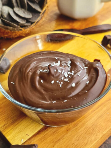 Crème dessert onctueuse au chocolat et lait de coco servie dans un bol en verre, saupoudrée de copeaux de noix de coco, présentée sur une planche en bois avec des carrés de chocolat noir et une coque de noix de coco remplie de pistoles de chocolat.
