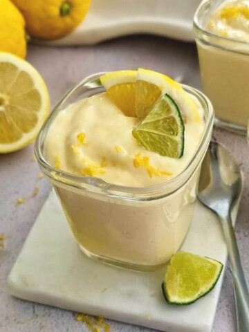 Trois verrines de mousse au citron onctueuse, décorées de quartiers de citron jaune et vert et de zestes frais, posé sur un support en marbre blanc avec des citrons entiers en arrière-plan.