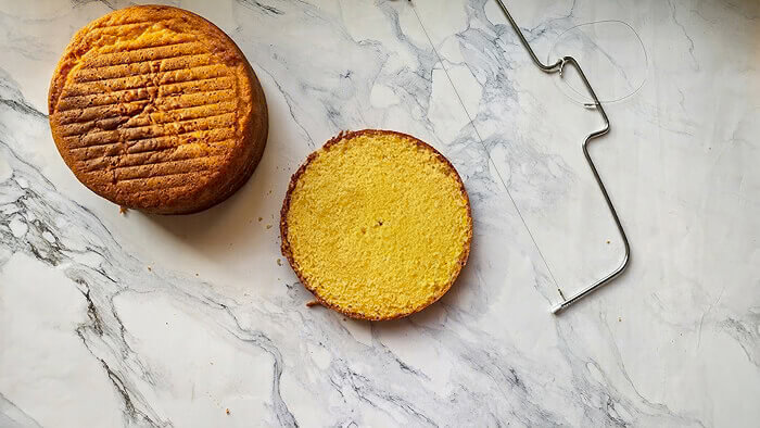 decoupe-tranche-molly-cake-avec-lyre Gâteau Molly Cake entier et une tranche parfaitement découpée côte à côte, avec un fil à découper (lyre) en métal sur un plan de travail en marbre, illustrant la technique de découpe.
