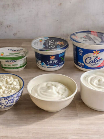 Une photo d'un plan de travail de cuisine alignant trois pots de produits laitiers de marques spécifiques (un petit Danone Cottage Cheese, un Skyr Siggi's moyen, un grand pot de Yoplait Calin Extra) et trois bols ouverts devant eux. Les bols révèlent la texture distinctive de chacun : à gauche, le cottage cheese grumeleux dans un bol bleu et blanc ; au centre, le skyr épais dans un bol beige ; à droite, le fromage blanc lisse dans un bol blanc.
