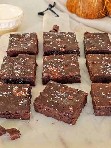 Présentation de neuf parts de fondant chocolat sans beurre crème de coco saupoudrées de noix de coco râpée sur une planche en marbre blanc. La mise en scène inclut une noix de coco entière et un bol de crème pour illustrer cette alternative saine au beurre.