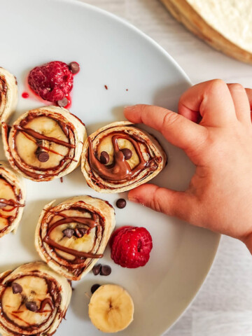 Vue d'ensemble d'une assiette blanche remplie de makis sucrés banane chocolat, entourée d'une banane entière, de pépites de chocolat et d'une pile de tortillas wraps.