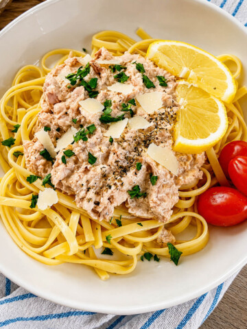 Une vue de dessus d'une assiette de pâtes linguines en sauce crémeuse au thon, richement garnie de copeaux de parmesan, de persil frais haché et d'herbes séchées. Le plat est présenté avec deux tranches de citron et quelques tomates cerises sur le côté, sur un fond texturé.