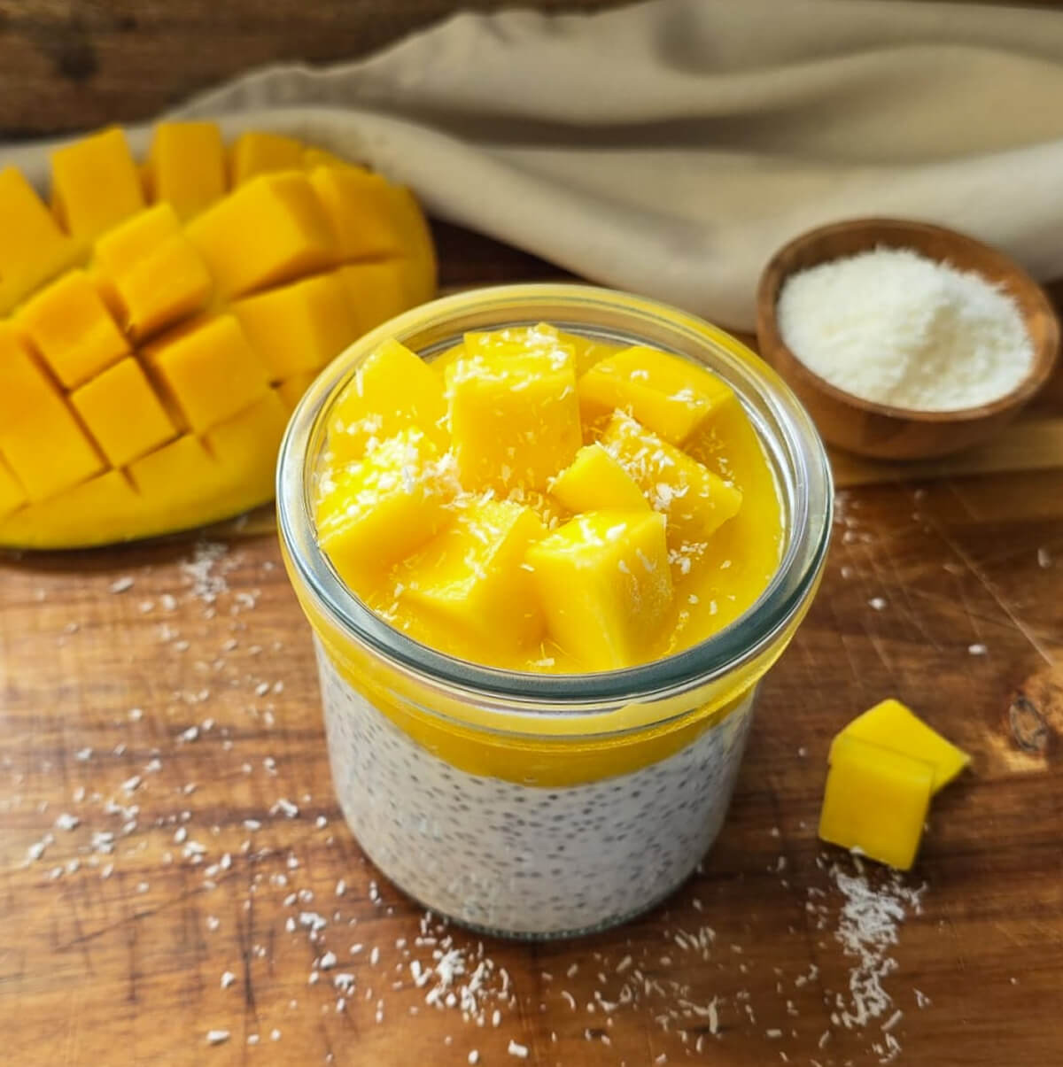 Pudding de chia au lait de coco dans un bocal en verre, surmonté d'une généreuse couche de coulis de mangue et de dés de mangue fraîche, saupoudré de noix de coco râpée.