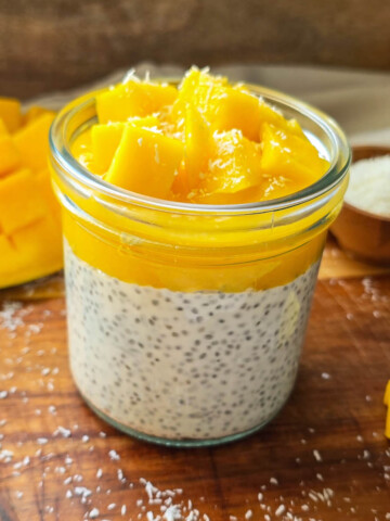 Gros plan d'un chia pudding mangue coco avec topping de mangue juteuse et noix de coco, accompagné d'une mangue découpée en damier en arrière-plan.