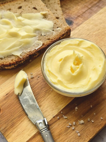 Beurre maison onctueux étalé sur une tranche de pain de campagne, avec un ramequin de beurre et un couteau Laguiole sur une planche en bois.