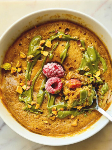 Vue de dessus d'un bowl cake à la pistache et framboise, au moelleux apparent, servi dans un bol blanc sur un marbre clair. Il est généreusement nappé d'une crème de pistache vert vif et parsemé d'éclats de pistaches grillées. Au centre, deux framboises surgelées sont entourées de baies cuites.