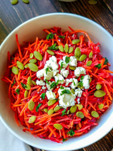 Un bol blanc rempli d'une salade de betterave, carotte et pomme, sur un fond en bois foncé, fourchette et torchon.