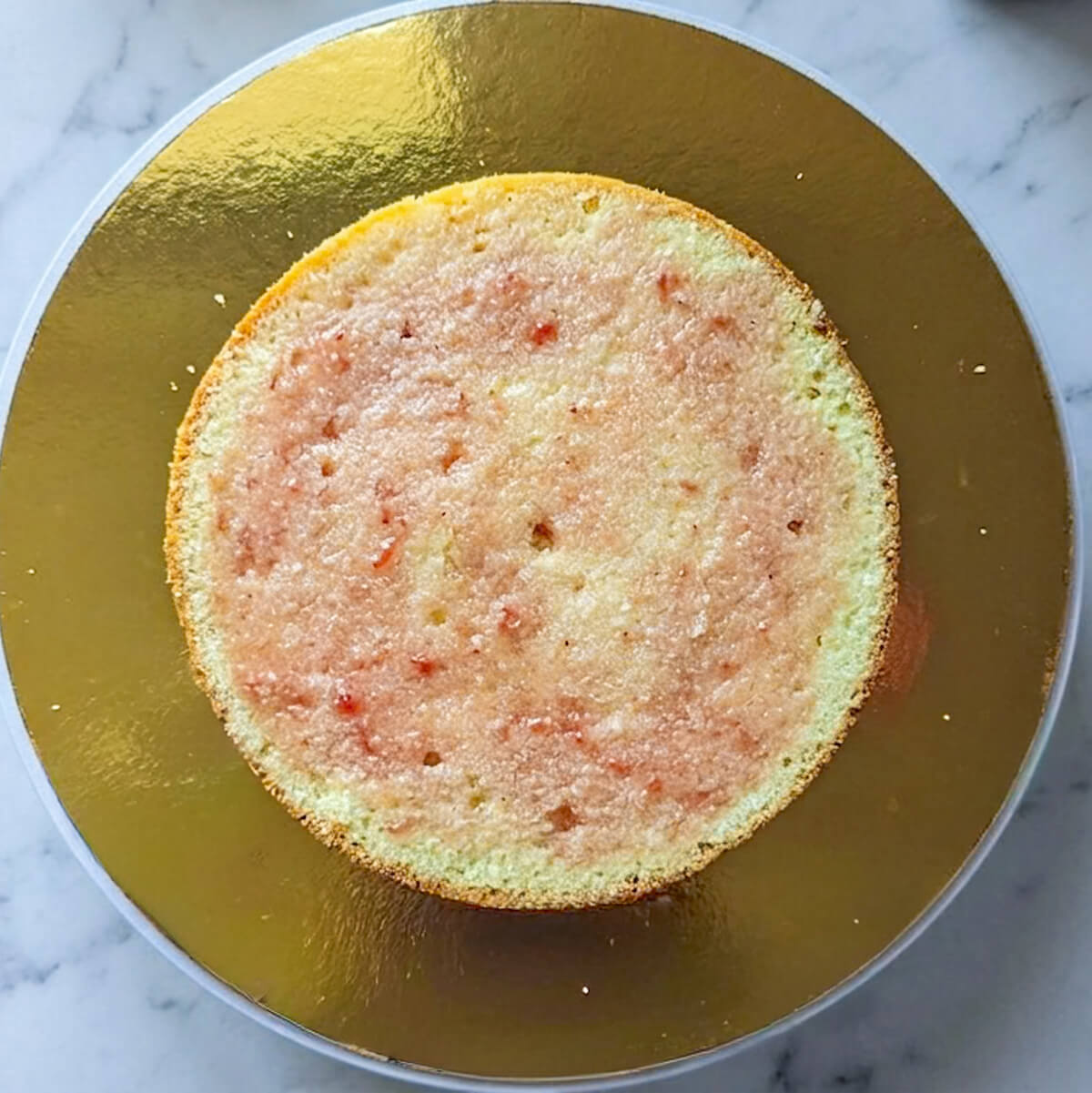 1-molly-cake-imbibé-layer-cake-fraise Vue de dessus d'un disque de Molly cake nature posé sur un support doré. Le biscuit est généreusement imbibé d'un sirop de fraises artisanal de couleur rose clair.