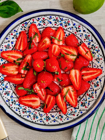 Une vue d'en haut d'une assiette en céramique décorée à motifs bleus, remplie de fraises fraîches et coupées en deux, garnies de rubans de basilic frais et de zestes de citron vert râpés. L'assiette est placée sur une table en bois clair, avec une fourchette à manche crème à gauche, des feuilles de basilic entières en haut à gauche, un citron vert entier en haut à droite, et un torchon à rayures vertes à droite.