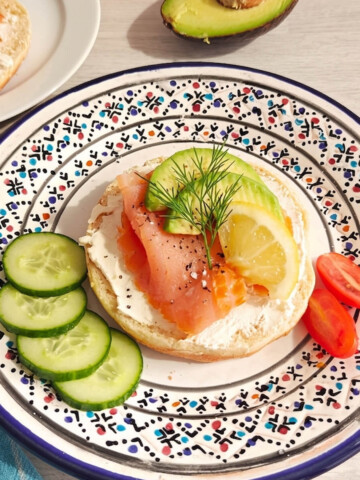 Un demi-bagel garni de fromage frais, d'une tranche généreuse de saumon fumé et de lamelles d'avocat. L'assiette est décorée d'une branche d'aneth, d'une tranche de citron, de rondelles de concombre et de tomates cerises.