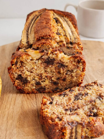 Un cake à la banane (banana bread) tranché sur une planche en bois, révélant une mie moelleuse aux pépites de chocolat fondu et éclats de noisettes, décoré d'une banane entière sur le dessus.