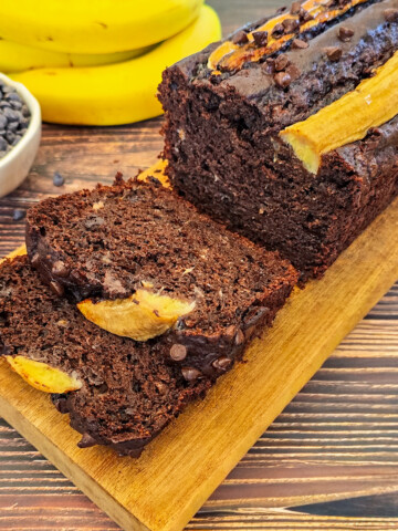 Gros plan sur des tranches de banana bread au chocolat sur une planche de bois, bananes et pépites de chocolat en arrière.