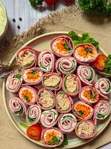 Gros plan sur une assiette blanche rectangulaire présentant trois bouchées de wraps à la betterave (makis). On distingue clairement une garniture au thon, une autre aux carottes râpées et une troisième au fromage frais. L'assiette est décorée d'une demi-tomate cerise et d'une feuille de chêne, avec une fourchette en argent gravée "Cuisine & Création". Le plateau principal est flou en arrière-plan.