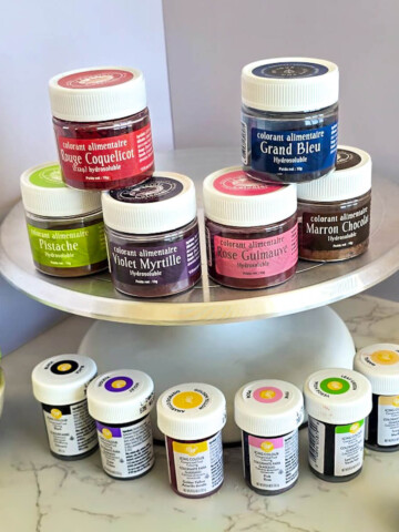 Une sélection de colorants alimentaires pour macarons : des pots de colorants hydrosolubles en poudre sur un support rotatif (rouge, bleu, violet, vert) et des petits flacons de colorant en gel Wilton au premier plan, à côté de macarons verts.