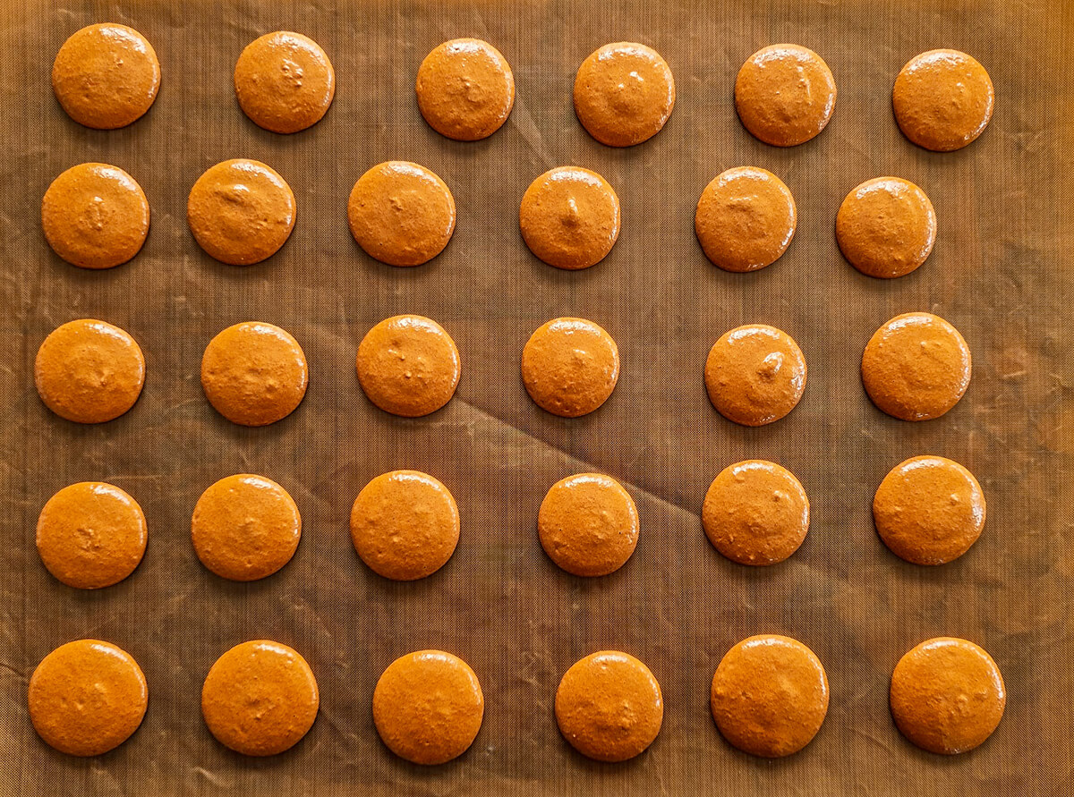 croutage-macarons-café Rangées de coques de macarons au marron clairs pochées sur une toile de cuisson en silicone marron, prêtes à être enfournées pour obtenir des biscuits lisses et parfaits.