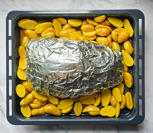 cuisson-gigot-agneau-pommes-terre Un gigot d'agneau dans du papier d'aluminium et des pommes de terre sur une plaque de cuisson.