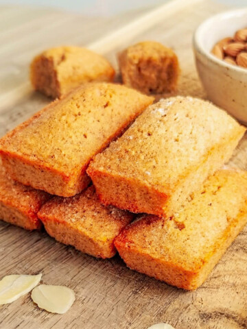 Gros plan sur une pile de financiers aux amandes dorés posés sur une planche en bois, avec un bol d'amandes entières en arrière-plan et des amandes effilées au premier plan.