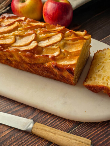 Gâteau au yaourt aux pommes moelleux présenté sur une planche en marbre avec des tranches découpées et deux pommes rouges en arrière-plan.
