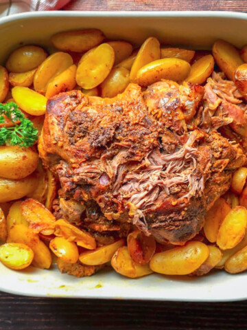 Gigot d'agneau rôti au four, effiloché et fondant, entouré de pommes de terre grenaille dorées dans un plat en céramique blanc.