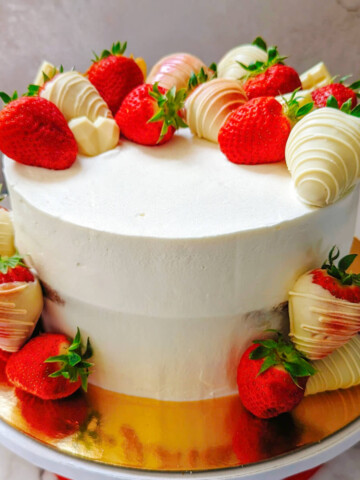 Un layer cake fraise mascarpone entier présenté sur un plat doré et marbré, avec un lissage parfait à la crème mascarpone et une décoration de fraises fraîches et chocolat blanc tout autour de la base et du dessus.