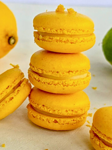 Une pile verticale de trois macarons jaunes au citron avec une collerette parfaite, entourée de citrons frais coupés, de zestes et de feuilles de basilic sur un fond blanc lumineux.