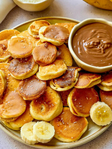 Une assiette remplie de "pancake cereal" (mini-billes de pancakes healthy à la banane) dorés et moelleux, parsemés de noix de coco râpée. Au centre, un bol de pâte à tartiner au chocolat pour le dip, avec des rondelles de banane fraîche et des pépites de chocolat en garniture.