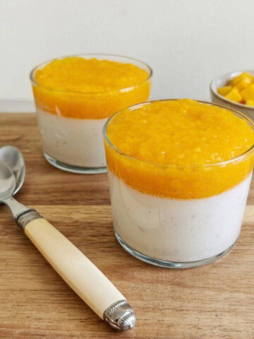 Deux verrines de dessert coco-mangue présentées côte à côte sur une planche en bois. Chaque verre montre des couches distinctes : une base de crème de coco blanche et un topping généreux de coulis de mangue jaune vif. À gauche, deux cuillères au manche ivoire et à droite, des feuilles de menthe fraîche et un bol de dés de mangue.