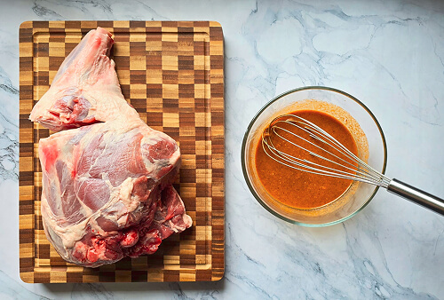 préparation-marinade-gigot-agneau-au-four-effiloché Gigot d'agneau cru sur une planche en bois à côté d'un bol de marinade orangée avec un fouet, sur un plan de travail en marbre.