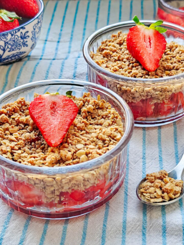 Deux verrines de crumble aux fraises décorées d'une demi-fraise, posées sur une nappe rayée à côté d'un bol de fruits frais.