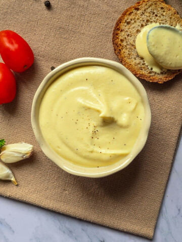 Vue de dessus d'un bol de mayonnaise œuf dur skyr sur un set de table en toile, entouré de deux tomates cerises, de gousses d'ail frais, d'un toast et d'une cuillère, sur un plan de travail en marbre.