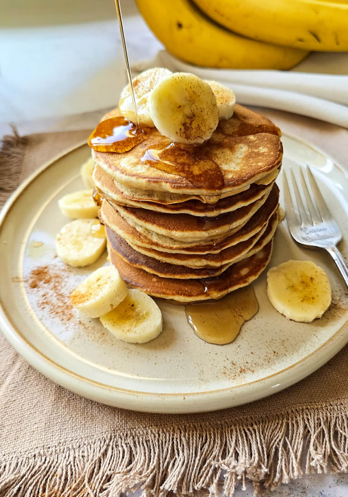 Haute pile de pancakes à la banane, miel doré et cannelle, servie avec des rondelles de banane fraîche sur une assiette en céramique, pour un petit-déjeuner gourmand.
